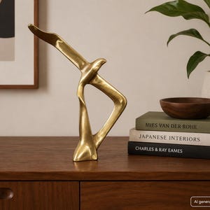 May include: A polished gold-toned metal sculpture depicting a stylized bird in flight with elongated, smooth, and curved forms. The abstract figure features a pointed beak, a slender neck, and a wide, sweeping wing structure that creates a hollow center.