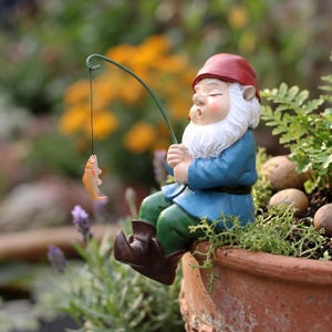 May include: A garden gnome figurine wearing a red hat, blue shirt, and green trousers, sitting on the edge of a terracotta pot. The gnome holds a thin green fishing rod with a black line and a small orange fish dangling from the end of the line.