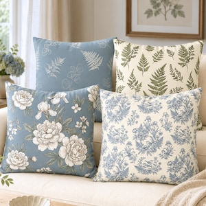 May include: Four square throw pillows arranged on a sofa. One is dusty blue with white floral patterns, one is dusty blue with white fern outlines, one is cream with green fern prints, and one is cream with a blue toile pattern of birds and flowers.