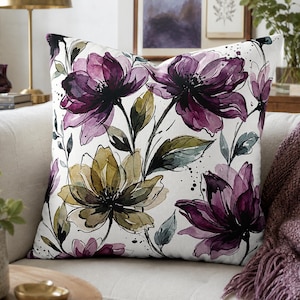 Puede incluir: Un cojín cuadrado con un estampado floral estilo acuarela sobre fondo blanco. El diseño muestra flores grandes de color morado intenso y verde oliva, con hojas y tallos de color verde oscuro y salpicaduras de tinta negra.