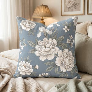 May include: A square throw pillow featuring a dusty blue background with a repeating pattern of large white peonies and smaller white blossoms with sage green leaves. The floral design is rendered in a detailed line art style with soft shading.