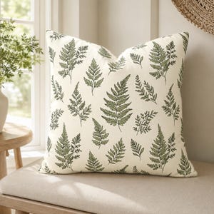May include: A square throw pillow featuring an off-white base color with a repeating pattern of green fern leaves printed across the front. The fabric has a visible woven texture, and the pillow is filled to maintain a plump, structured shape.