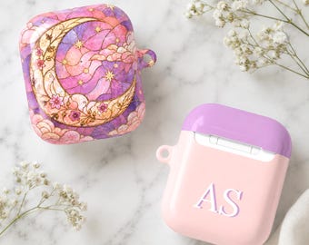 May include: Two protective cases for wireless earbuds. One case features a purple and pink celestial design with a golden crescent moon, stars, and clouds. The second case is light pink with a solid purple lid and the white letters AS printed.