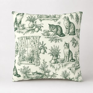 May include: A square throw pillow featuring a cream-colored fabric with a dark green toile-style print. The pattern depicts multiple cats, potted plants, lemons, teacups, and a window with curtains, creating a detailed, repeating illustrative scene.