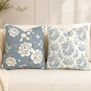 May include: Two square throw pillows. The left pillow features a dusty blue background with large white peony flowers and green leaves. The right pillow has a cream background with a blue toile pattern of small birds, flowers, and foliage.