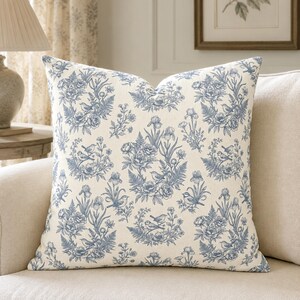 May include: A square throw pillow featuring a cream-colored fabric base with a repeating blue toile pattern. The design displays intricate illustrations of small birds perched among clusters of flowers, ferns, and iris blossoms in a classic style.