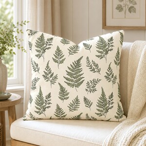 May include: A square throw pillow featuring an off-white base color with a repeating pattern of dark green fern leaves. The botanical print covers the entire front surface of the pillow, showcasing various sizes and orientations of fern fronds.