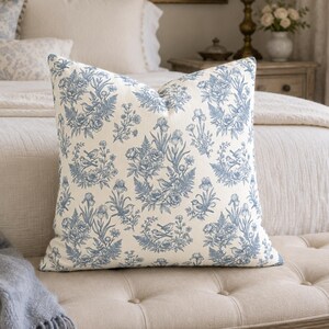 May include: A square throw pillow featuring a cream-colored fabric base with a repeating blue botanical print. The pattern consists of detailed illustrations of small birds perched among various flowers, leaves, and ferns in a toile-style design.