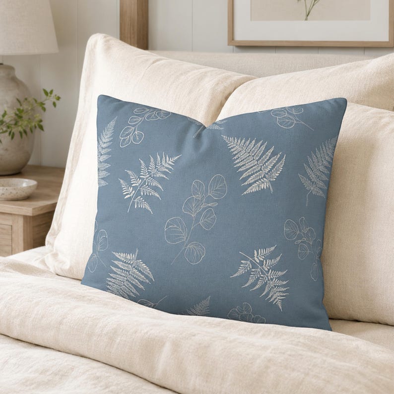 May include: A square throw pillow featuring a dusty blue fabric base with a repeating white line art pattern of fern fronds and eucalyptus leaves. The design is printed across the front surface of the pillow, which has a smooth, matte texture.