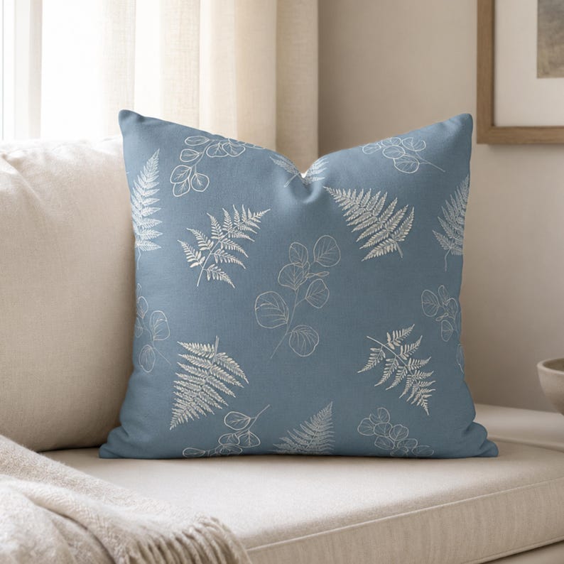May include: A square throw pillow in a dusty blue color featuring a repeating pattern of white line-drawn botanical elements, including various fern fronds and eucalyptus-style leaves scattered across the front surface of the fabric cover.