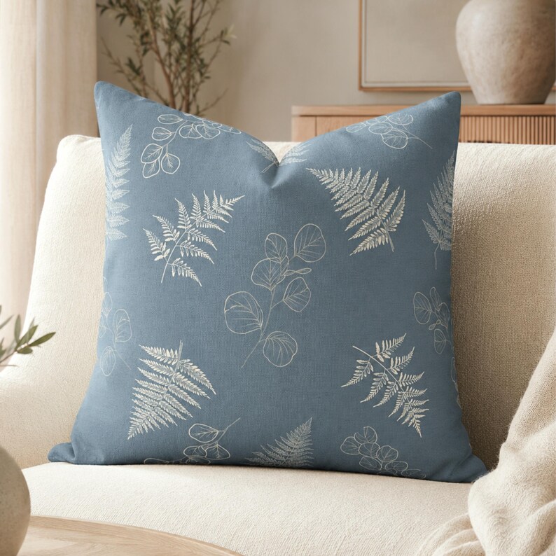 May include: A square throw pillow in a dusty blue color featuring a repeating white line art pattern of fern fronds and eucalyptus leaves. The fabric has a visible woven texture, and the design is printed across the entire front surface.