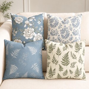 May include: Four square throw pillows featuring distinct botanical patterns. One pillow has white floral illustrations on a blue background, another has blue toile-style floral prints, one has white fern and leaf outlines on blue, and one has green ferns on cream.