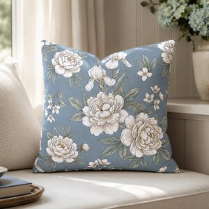 May include: A square throw pillow featuring a dusty blue background with a repeating pattern of white and cream-colored peonies, iris flowers, and small blossoms, accented by sage green leaves and stems throughout the fabric design.