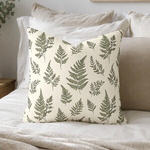 May include: A square throw pillow featuring a cream-colored background with a repeating pattern of green fern leaves. The botanical illustration style uses dark green outlines and muted green fill colors to depict various sizes of fern fronds.