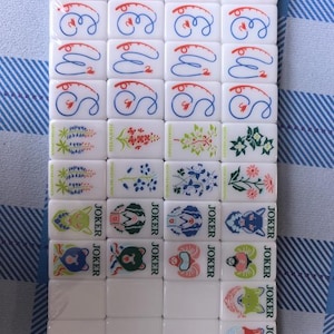 Nature-inspired American mahjong tile set with glossy white acrylic tiles featuring blue fish dot tiles, green bamboo designs, coral-inspired crack tiles, illustrated wildflower flower tiles, and colorful animal joker tiles. Modern engraved NMJL-style mahjong set arranged in clean flat lays with minimalist styling and outdoor mountain aesthetic, photographed on a soft white background with premium collectible game design.