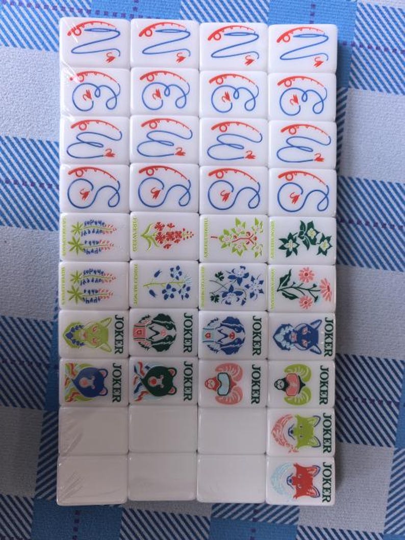 Nature-inspired American mahjong tile set with glossy white acrylic tiles featuring blue fish dot tiles, green bamboo designs, coral-inspired crack tiles, illustrated wildflower flower tiles, and colorful animal joker tiles. Modern engraved NMJL-style mahjong set arranged in clean flat lays with minimalist styling and outdoor mountain aesthetic, photographed on a soft white background with premium collectible game design.