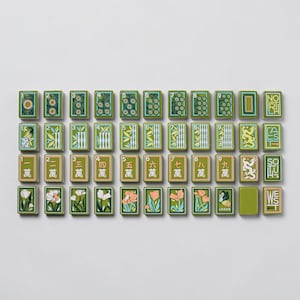 Könnte beinhalten: Eine Anordnung von grünen und cremefarbenen Mahjong-Steinen auf weißem Grund. Die Spielsteine zeigen verschiedene Designs, darunter Blumen, Bambusstängel und chinesische Schriftzeichen. Einige Steine sind nummeriert.