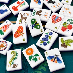 May include: A collection of white Mahjong tiles with colorful illustrations. Each tile features a unique design, including butterflies, flowers, and berries. The tiles have blue edges and are arranged on a green surface.