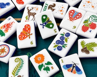 Könnte beinhalten: Eine Sammlung weißer Mahjong-Steine mit farbenfrohen Illustrationen. Jeder Stein zeigt ein einzigartiges Design, darunter Schmetterlinge, Blumen und Beeren. Die Steine haben blaue Ränder und sind auf einer grünen Oberfläche angeordnet.
