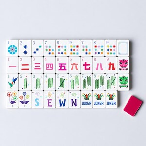 以下が含まれることがあります： カラフルなシンボルと数字が描かれた白い麻雀牌のセット。花、鳥、漢字など、さまざまなデザインが施されています。小さなピンク色の牌が横に置かれています。「SEWN」の文字が1つの牌に表示されています。