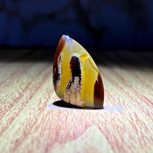 Puede incluir: Una piedra preciosa pulida en forma de lágrima con una superficie lisa que presenta bandas distintas y arremolinadas de color marrón caoba profundo, amarillo dorado translúcido y blanco crema. La piedra tiene un acabado brillante.