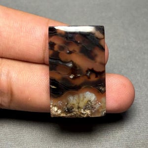 May include: A rectangular polished gemstone cabochon featuring a smooth surface with swirling patterns of deep black, translucent brown, and cream-colored inclusions, with a textured, crystalline cluster at the base.