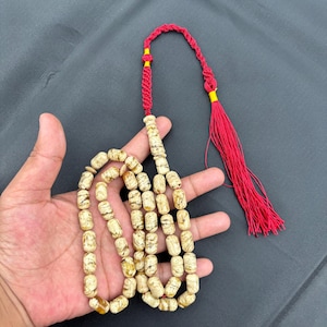 May include: A strand of prayer beads with off-white, cylindrical beads featuring brown marbling. The beads are strung on a red cord, which ends in a red tassel. The cord is accented with yellow thread.