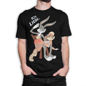 May include: A black short-sleeved t-shirt featuring a graphic of two cartoon rabbits. One rabbit is standing behind the other, who is bent over a chair. The text "ICH LIEBE..." is printed in white above the characters in a bold, sans-serif font.