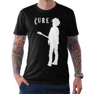May include: A black short-sleeved t-shirt featuring a white graphic print of a person with spiky hair holding a guitar. Above the figure, the text reads THE CURE in a stylized, distressed white font. The shirt has a crew neckline and a straight hem.