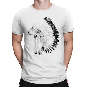 Peut inclure: Un t-shirt blanc à manches courtes arborant un grand imprimé graphique noir et blanc centré représentant une coiffe à plumes traditionnelle. Le motif présente des détails complexes de plumes individuelles et une bande décorative contrastée.