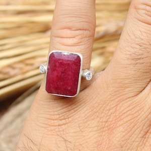 May include: A silver ring featuring a large, rectangular, deep red gemstone. Two smaller, clear stones flank the main stone. The ring is displayed on a hand, with a background of wheat and rope.