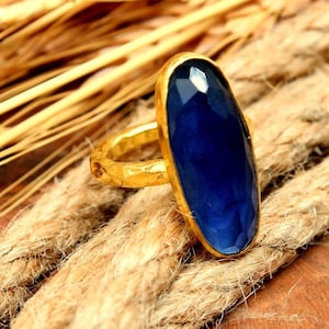 May include: A gold-toned ring with a large, oval, deep blue gemstone. The ring is set against a backdrop of thick, textured rope and wheat stalks, creating a rustic aesthetic. The gemstone is approximately 2.5 cm in length.