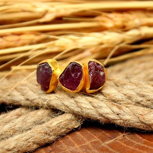 May include: A gold ring with three raw, deep red gemstones. The ring is set on a thick, textured rope, with a background of wheat stalks. The ring's design is simple, highlighting the natural beauty of the stones.