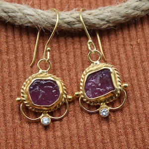 May include: A pair of gold earrings featuring a deep pink gemstone. The earrings have a decorative gold border and a small, clear stone at the bottom. The earrings are displayed on a brown surface.