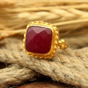 May include: A gold-colored ring with a square, faceted, deep red gemstone. The ring has a textured, rope-like border around the stone and a floral design on the side. The ring is displayed on a textured, brown rope.