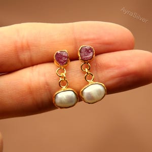 May include: Gold-toned earrings featuring a small, raw ruby at the top and a white, oval-shaped pearl at the bottom. The earrings are connected by a series of small gold loops. The earrings are held up by a person's fingers.
