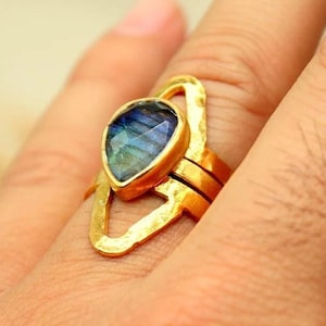 May include: A gold-colored ring with a teardrop-shaped, blue and green gemstone. The ring has a unique open design and is worn on a finger. The ring is a piece of jewelry.
