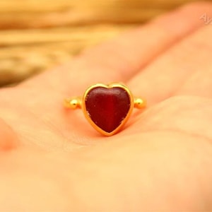 Pode incluir: Um anel dourado com uma pedra vermelha escura em forma de coração. O anel é segurado em uma mão, com o logotipo AyraSilver visível. O anel tem um acabamento liso e polido. A pedra tem cerca de 1 cm de largura.