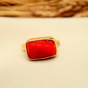 May include: A gold-toned ring with a rectangular, vibrant red stone. The ring is set on a cream-colored fabric, with a blurred background of dried plants. The AyraSilver logo is visible in the top right corner.
