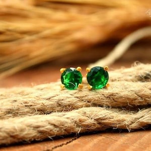 Può includere: Un paio di orecchini a bottone color oro, ciascuno con una gemma verde rotonda e vibrante. Gli orecchini sono posizionati su una superficie strutturata color marrone chiaro, creando un contrasto con le pietre verde brillante. Gioielli.
