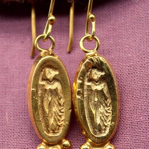 May include: Gold-toned earrings with an oval pendant featuring a carved figure. The earrings have a detailed design with a three-bead cluster at the bottom. The background is a muted purple fabric.