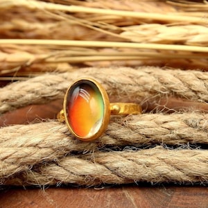 May include: A gold-colored ring with an oval-shaped, color-changing stone. The stone transitions from orange to yellow to green. The ring is set on a thick, textured rope, with wheat stalks in the background.