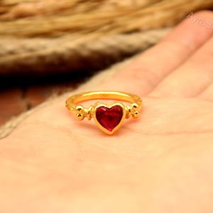 Puede incluir: Un anillo de tono dorado con una piedra roja central en forma de corazón. La banda tiene un diseño decorativo y texturizado a ambos lados del engaste del corazón. El texto AyraSilver es visible en la esquina superior derecha.