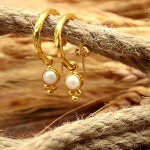 May include: Gold-coloured hoop earrings with a hammered texture. Each earring features a dangling pearl and three small gold beads. The earrings are displayed on a thick, textured rope against a blurred background of wheat.