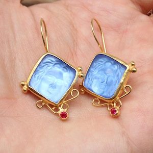 May include: A pair of gold-toned earrings with square, light blue cameo-style pendants. The pendants feature a classical figure. Each earring has a red gemstone and decorative gold filigree.