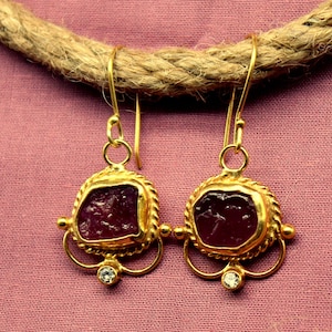 Può includere: Un paio di orecchini color oro con una pietra rosso scuro al centro. Gli orecchini hanno un bordo dorato decorativo e una piccola gemma trasparente in basso. Gli orecchini sono appesi a una corda.