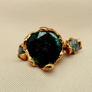 May include: A gold-toned ring with a large, faceted, dark green gemstone at the center. Two smaller, gray-toned stones flank the central stone. The ring has a textured, organic design.
