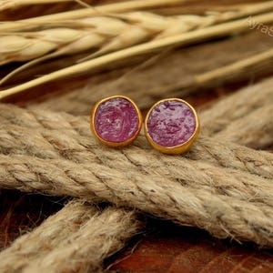 May include: A pair of gold-colored stud earrings with a round, textured, magenta-colored stone set in each. The earrings are displayed on a textured, brown rope surface with wheat stalks in the background.