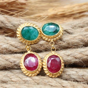 May include: A pair of gold-colored earrings with emerald green and ruby red gemstones. The earrings have an oval shape with a textured gold border. The gemstones are set in a gold bezel setting, and the earrings are displayed on a textured rope surface.