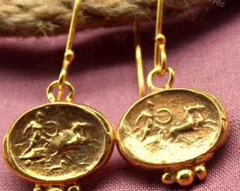 May include: A pair of gold-colored earrings with an oval pendant featuring a detailed relief design. The earrings have a hook closure and small, decorative beads at the bottom of the pendant. The background is a muted pink.